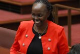 Lucy Gichuhi, wearing a black top and red jacket, smiles in the Senate. She is wearing a silver pendant around her neck.