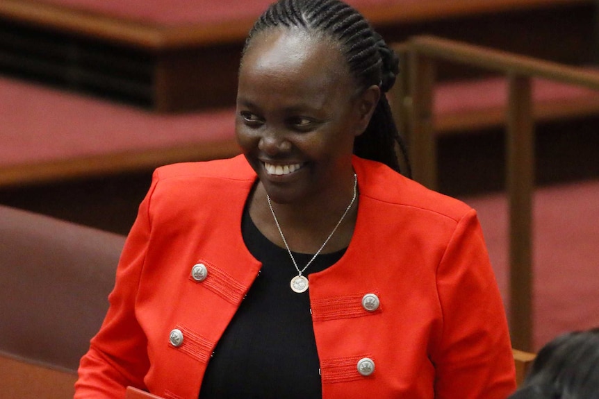 Lucy Gichuhi, wearing a black top and red jacket, smiles in the Senate. She is wearing a silver pendant around her neck.