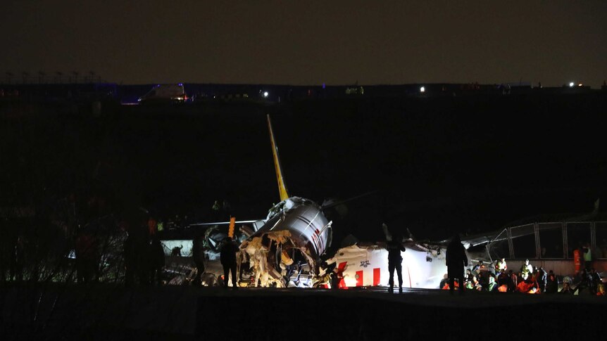 Lights show parts of a plane wreck with silhouettes of people standing around on runway.