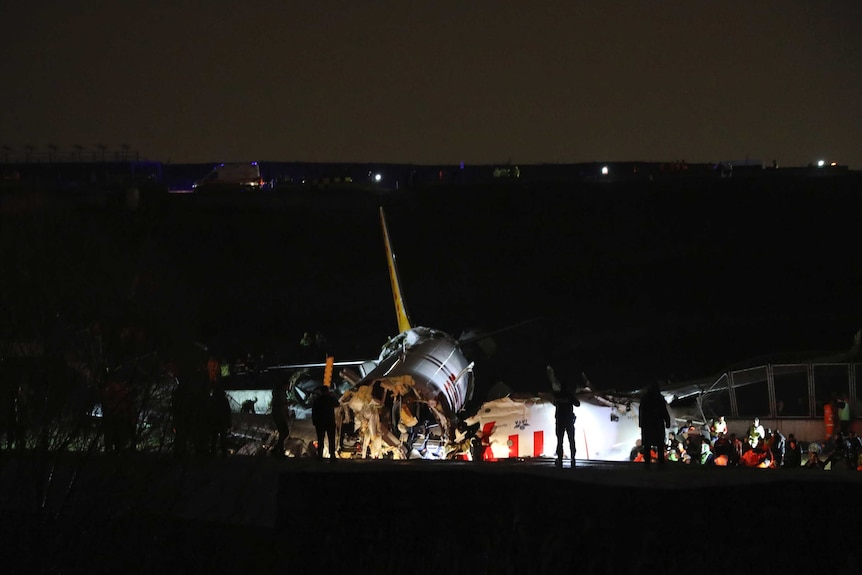 Lights show parts of a plane wreck with silhouettes of people standing around on runway.