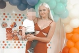 A young women with long blonde hair holds her child in front of balloons