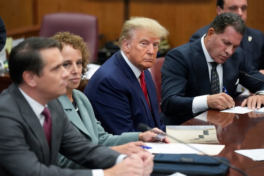 Donald Trump sitting at a bar table, flanked by lawyers