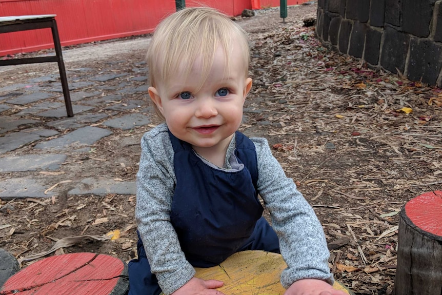 A blonde toddler holds himself up on a log and smiles at the camera