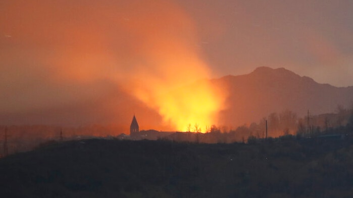 You see a church steeple silhouette lit by orange flames at night seen in the distance behind green hills.