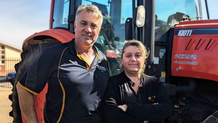 A grey-haired man stands hands next to a woman who has her arms crossed in front of a tractor.