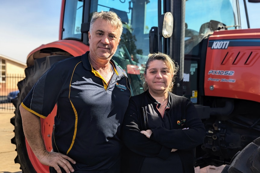 A grey-haired man stands hands next to a woman who has her arms crossed in front of a tractor.
