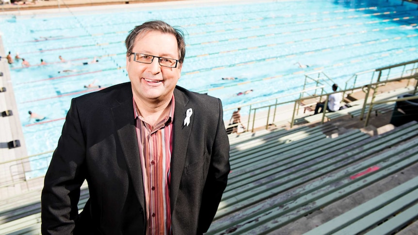 A man wearing glasses and a dark jacket standing in the bleachers with a swimming pool in the background.