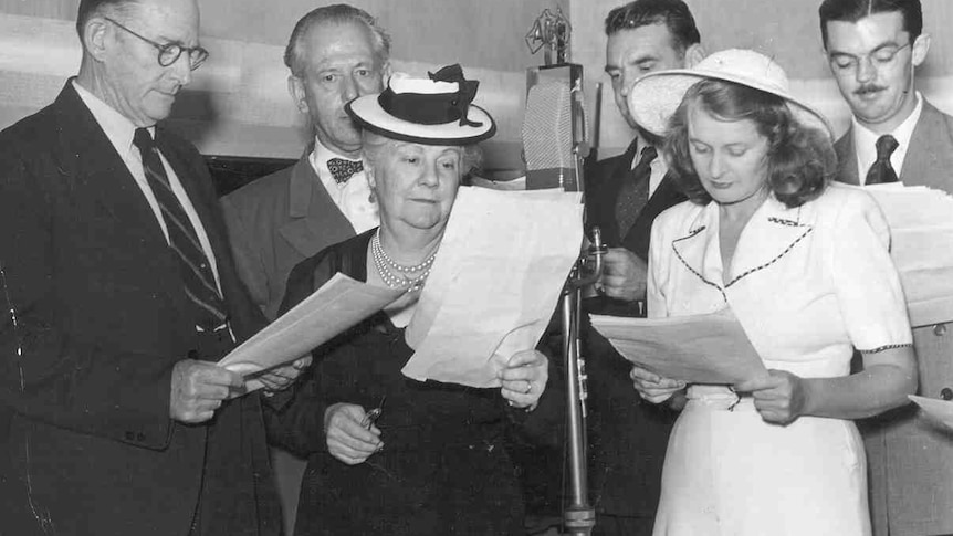 A black and white photograph of two well dressed women holding scripts in front of a microphone with four men with scripts behi