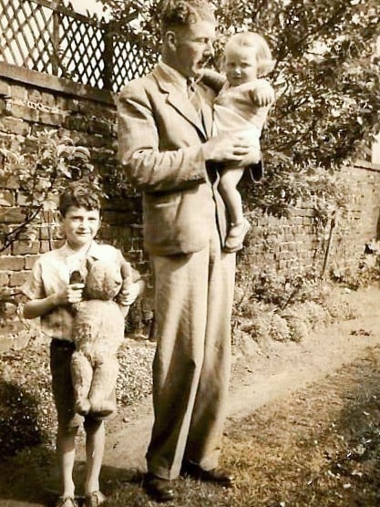 A black and white photograph shows an older gentleman holding a toddler aged girl and a young john standing beside them