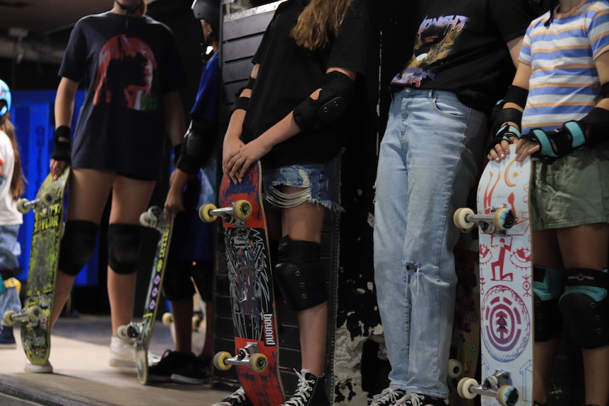Girls wait their turn, ready to drop in. They're all holding their skateboards, wearing knee and elbow pads.