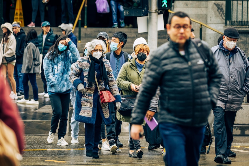 A group of pedestrians, all rugged up and several wearing masks, cross Flinders Street on a wet day.