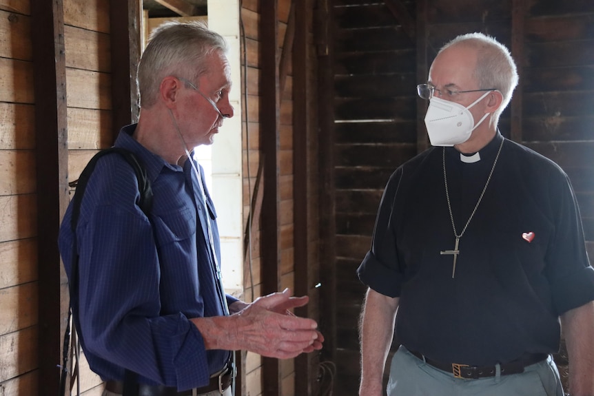 A man with a blue shirt and oxygen tubes stands talking with another man in a black shirt and clerical collar.