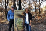A man and a woman standing near a sign with burnt-out bushland in the background.