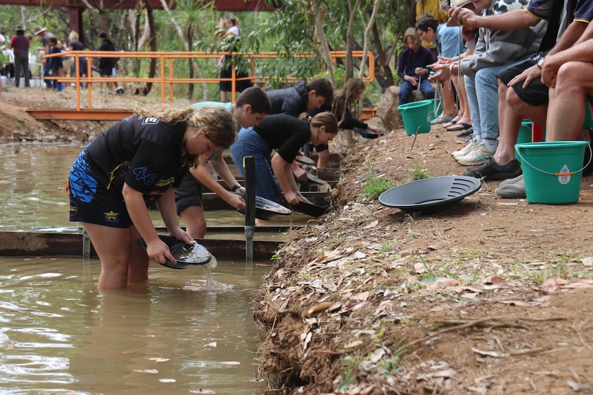 A group of people pan for gold in a river bed as people watch on along the river bank.