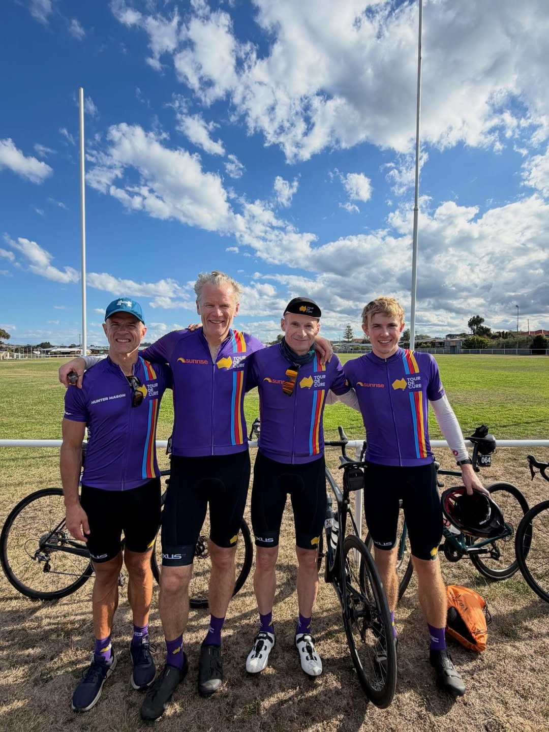 Four men stand arm in arm wearing matching purple cycling gear, their bikes behind them. They stand at football field, blue sky