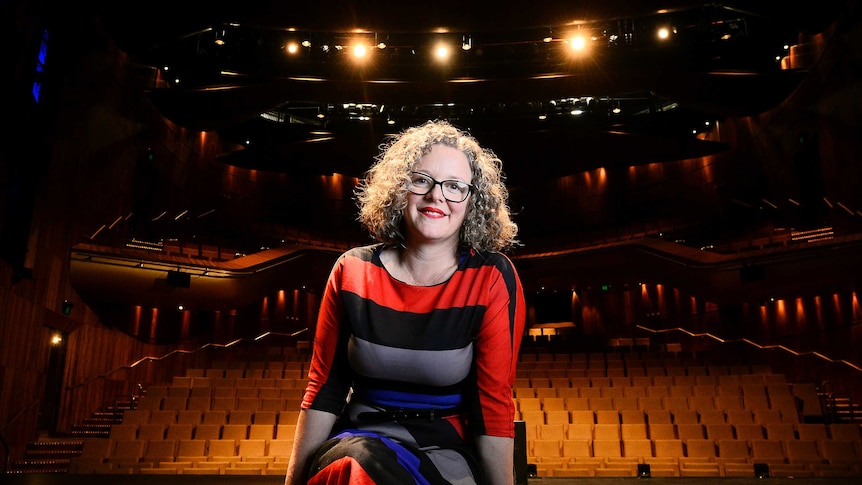 A woman with curly hair and glasses sit smiling on a stage with an empty rows of seats in the background