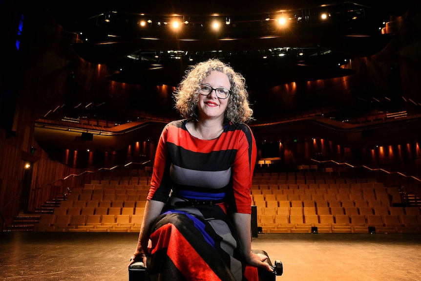 A woman with curly hair and glasses sit smiling on a stage with an empty rows of seats in the background