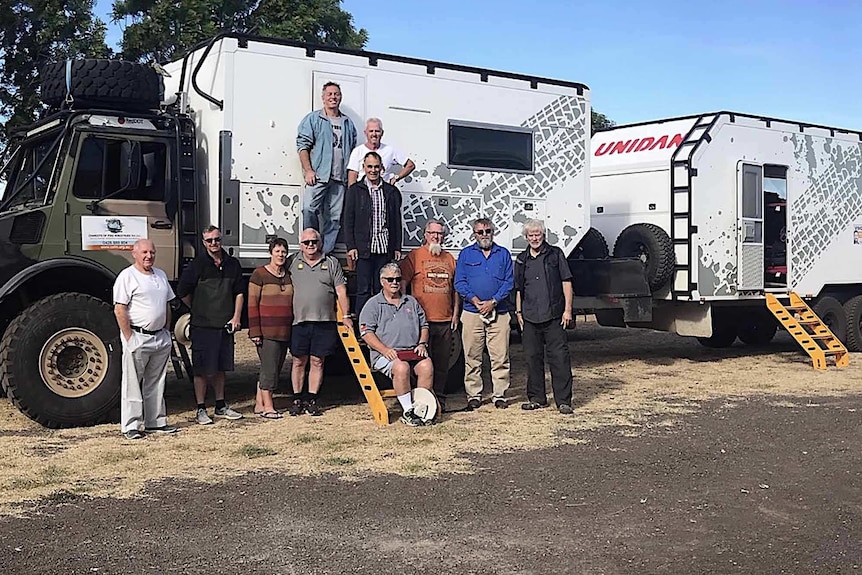 A group of white men and women stand in front of a large all-terrain truck and trailer.