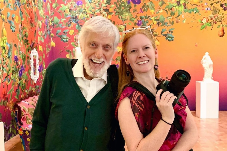 An elderly man and a woman holding a camera, smile for a photo together.