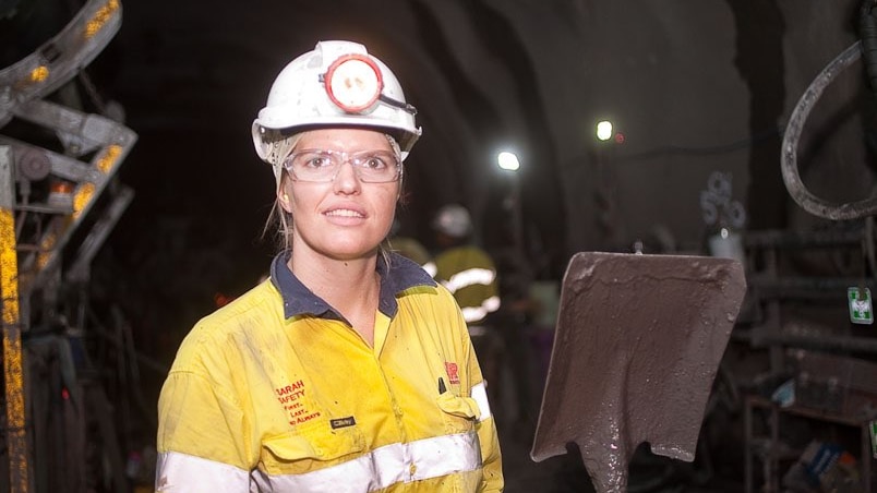 A woman with a hard hat on and high-vis shirt is in an underground environment smiling at the camera