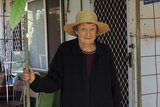 An old woman with a straw hat stands in front of her front door.