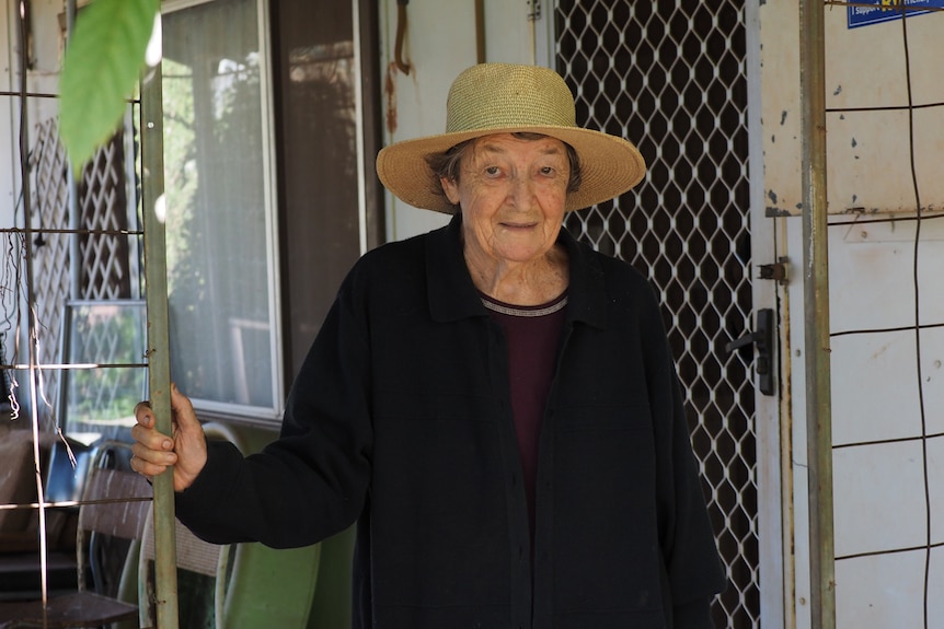 A mature woman with a straw hat stands in front of her front door.