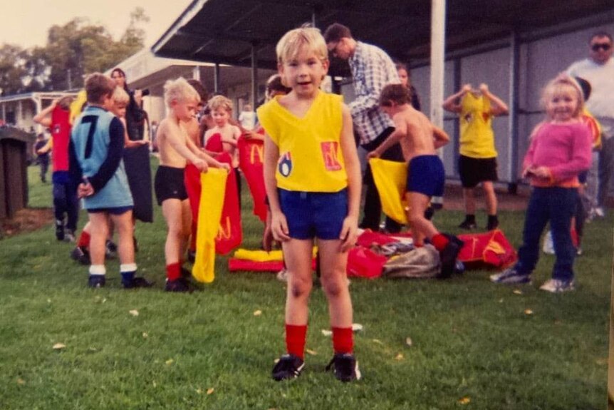 A young boy wears a footy uniform with a group of kids behind him.
