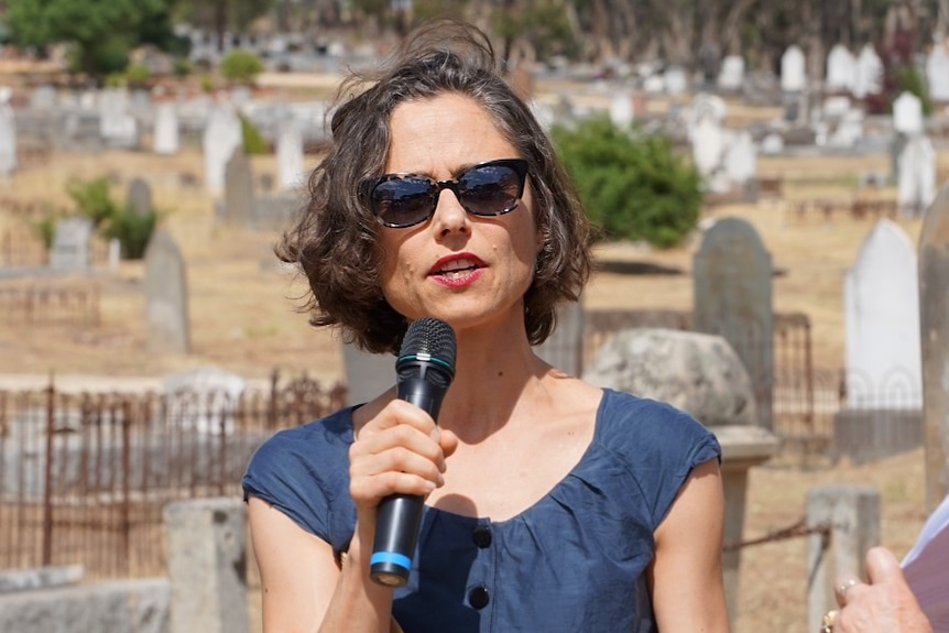A woman wearing sunglasses speaks into a microphone at a cemetery.