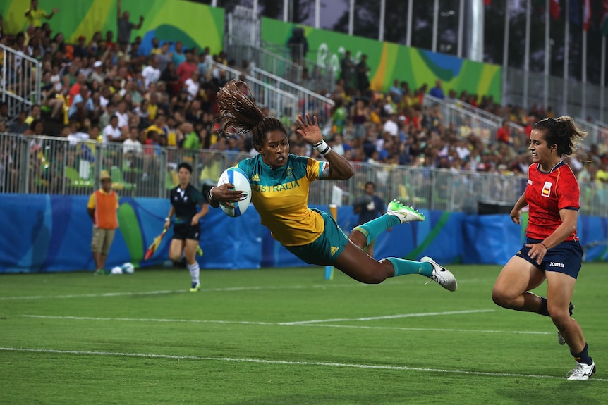 An Australian rugby sevens player flies through the air over the try-line to score with the referee standing to one side. 