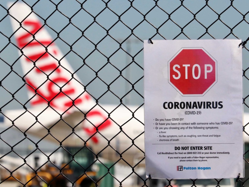 A coronavirus warning sign on a wire fence, in front of a Virgin plane