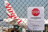 A coronavirus warning sign on a wire fence, in front of a Virgin plane