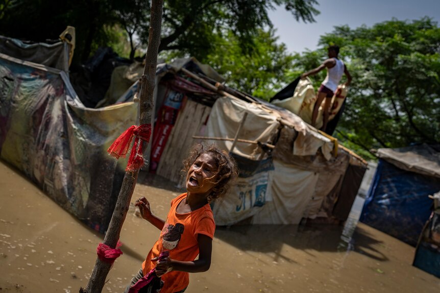 A child cries as she stands in floodwater. Behind her a man pulls apart a tent inundated.