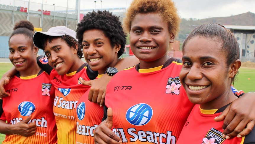 Five Papua New Guinean women rugby league players in red shirts embrace and smile for the camera.