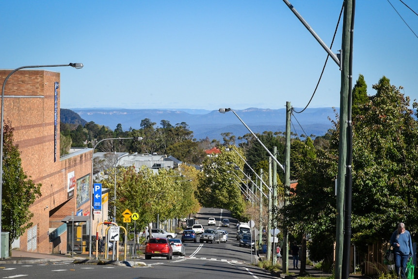 A street dips downhill with a large red brick building to the left and escarpments and mountains in the far distance.