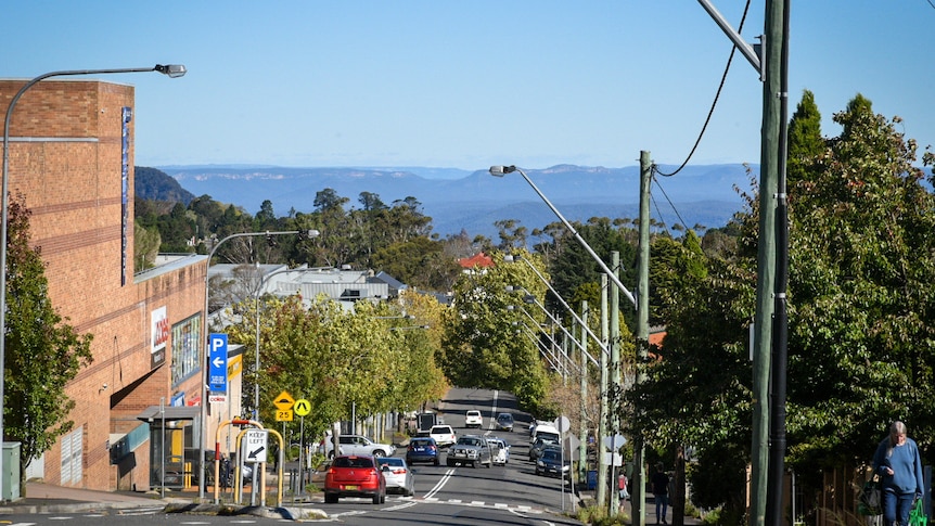 A street dips downhill with a large red brick building to the left and escarpments and mountains in the far distance.