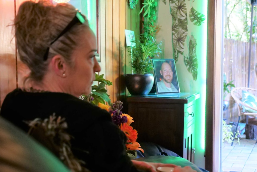 A woman sits on a couch in front of her husband's portrait.