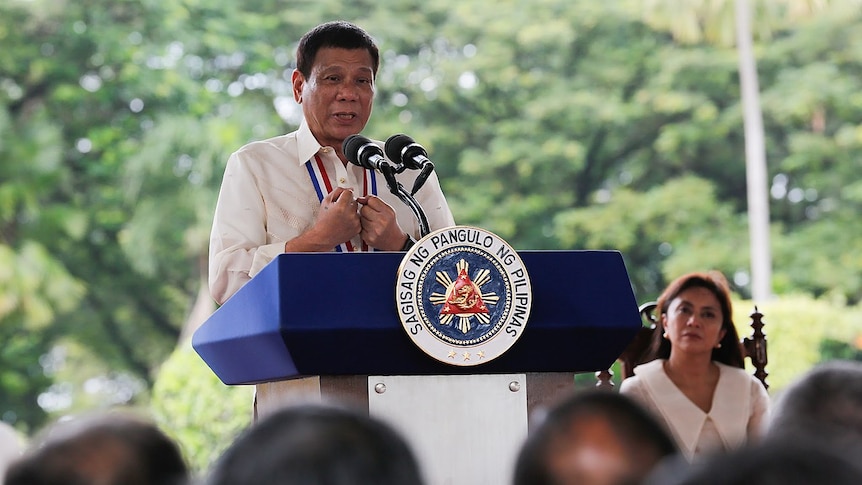 Rodrigo Duterte stands behind a large blue lectern carrying the Filipino presidential seal in front of greenery.