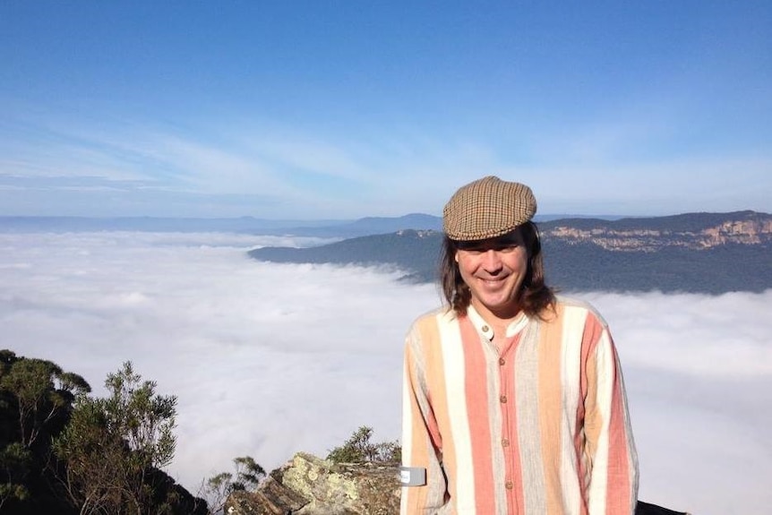 a man in an orang striped shirt and beret stands in front of a valley filled with clouds and a sandstone escarpment behind it.