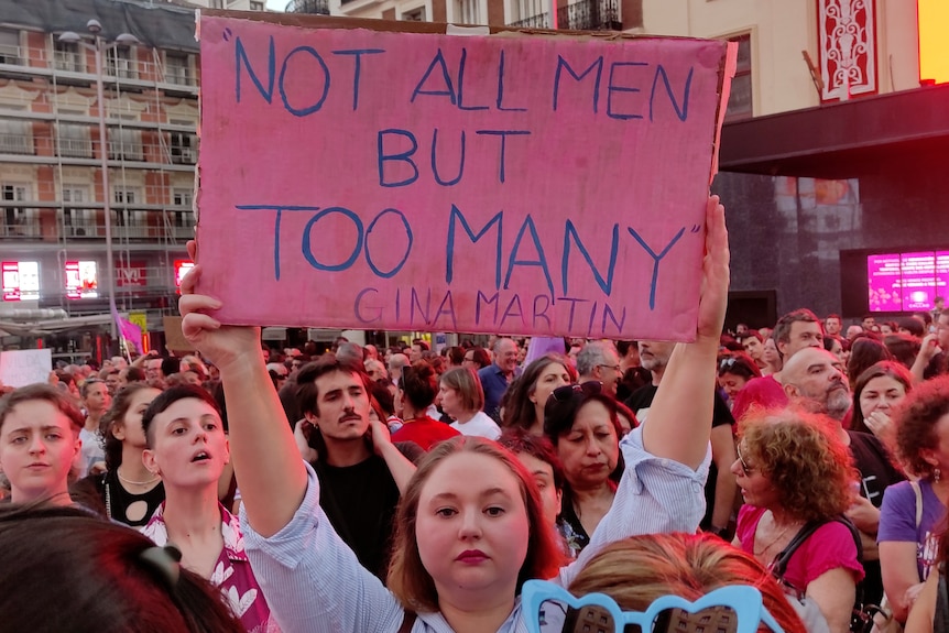 A woman in a sea of protesters holds a placard that reads "Not all men but too many"