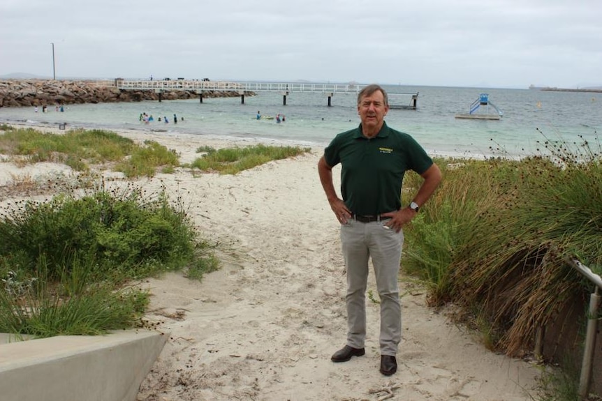 A man in a polo shirt stands on a beach with his hands on his hips.