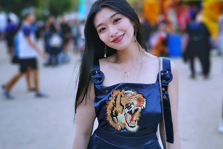 A young woman in a black dress embroidered with the face of a tiger, smiles at a fairground.