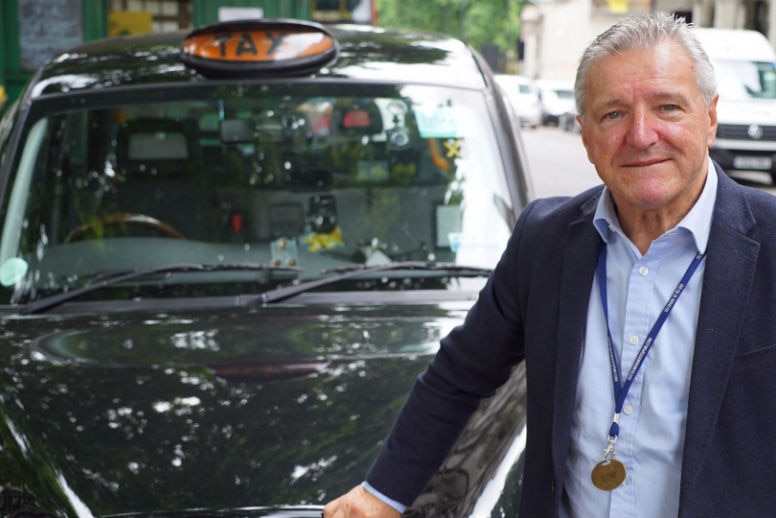 A man in a sports coat and business shirt wears a medal around his neck as he leans against a black cab on a London street.