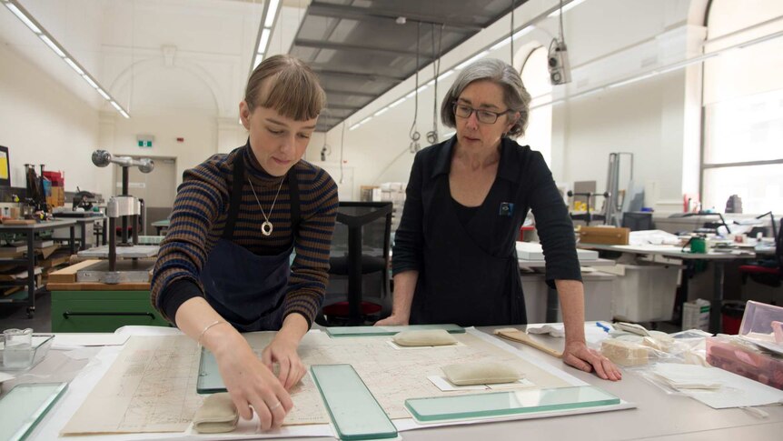 Two women look at a map, glass and weights hold down the paper. Book presses in background.