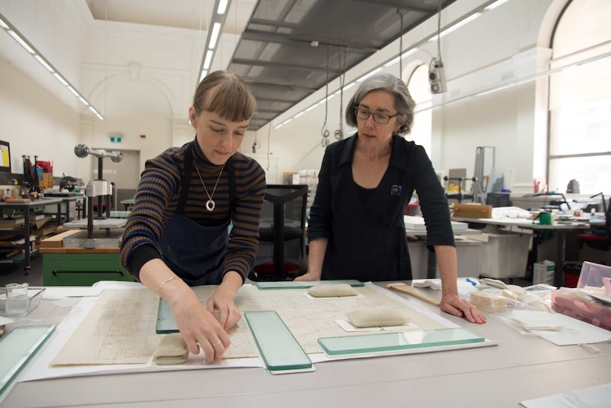 Two women look at a map, glass and weights hold down the paper. Book presses in background.