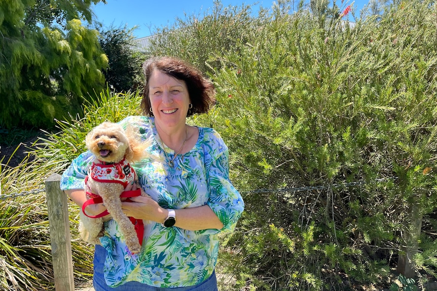 Woman wearing blue-and-green top stands in garden holding a small brown dog in a harness and smiling.
