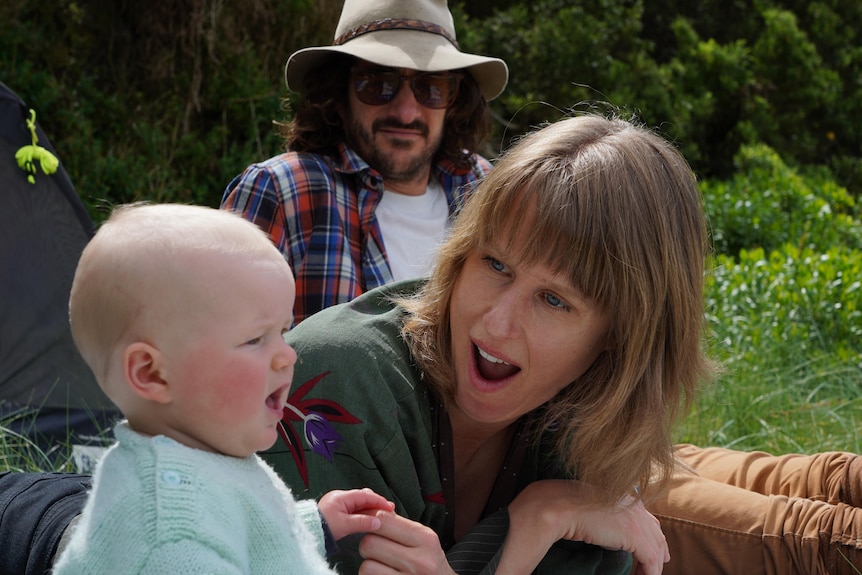 A woman lies on her stomach with an animated face to entertain a baby, a man is in the background