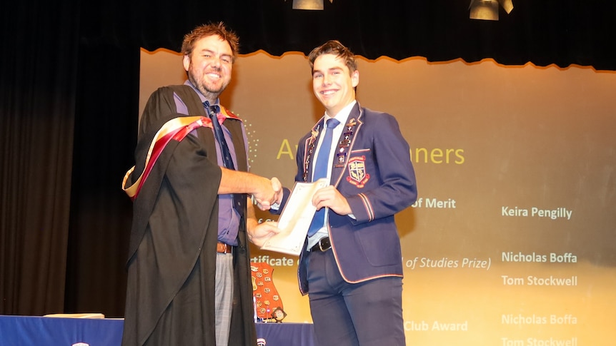 A young man in school uniform receives an award from a man in an academic gown.