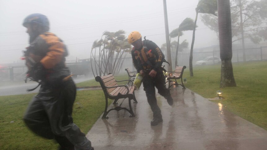 Two figures in rescue uniforms run through the rain in a park as palms behind them bend in the high winds.