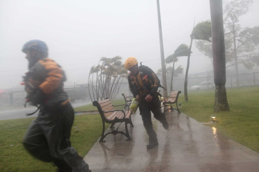 Two figures in rescue uniforms run through the rain in a park as palms behind them bend in the high winds.