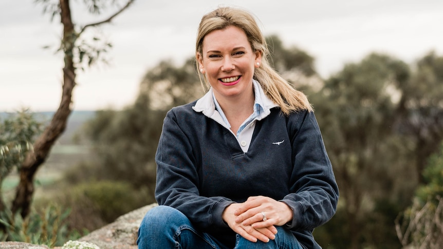 Kate Gunn, a smiling blonde-haired woman wearing a navy jumper and jeans, sits on a rock with gum trees behind her. 